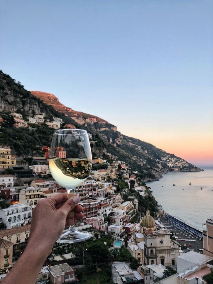 A glass of white wine raised over Positano at dusk, houses stacked above the sea